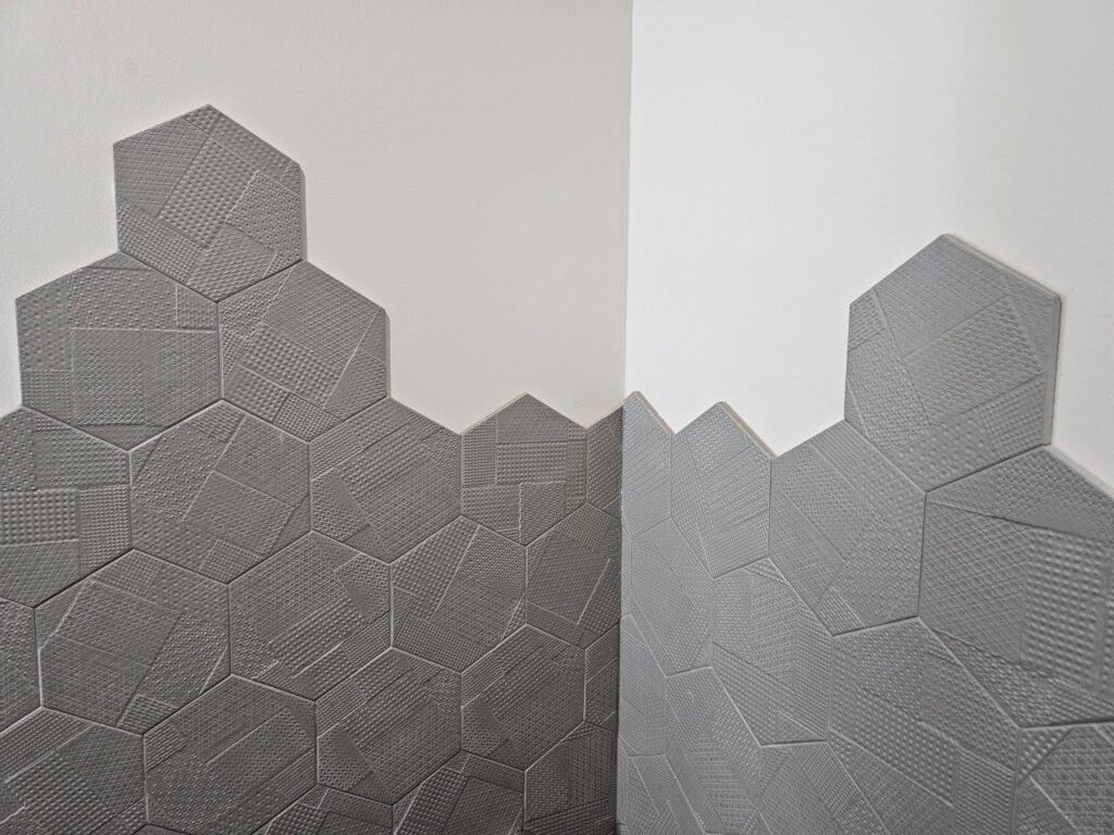 The tile used on his bathroom is an unterminated or hexagonal tile with an embossed texture. Unlike most wall tile installations which terminate all the way up at the corner of the joint between the wall plain or have a termination strip, this tile is installed with a raw staggered edge. Sometimes an edge like this is called a floating edge. If you look in the picture below, you can see that the tile at the vertical inside corner of the wall is cut to fit the shape provided by the wall plane. The top of the tile though is left raw to just follow a staggered edge of the hexagonal tile itself. The staggered unterminated edge of the tile doesn't necessarily look bad, in this example. They've been careful during installation to not let mortar ooze out in a way that looks rough or unfinished. At the lower tiles though, in the picture below, you can see the top of that unterminated edge. There are two problems with that kind of unterminated edge. First, you can see the raw edge of the tile itself. Since this is not a solid piece of natural material, the front or exposed face of the tile is a different color than the internal composition or body of the tile. When it's exposed like this and doesn't have a terminating edge you can see that difference which makes it visually apparent that it's not a real material. It's a manufactured material. The second problem with this type of unterminated edge is that the mortar itself has a thickness of roughly 1/8 of an inch. When applied with a 3/16 inch Notch trowel, the mortar becomes compressed but still even when flattened as much as possible there still is a thickness of that mortar. Once it is dry and cured, you can still see the thickness of that mortar. In almost all cases of tile installation, unlike stone masonry or even brick masonry, the tile mortar is not intended to be exposed at the joints. In between joints, in the field of the tile, the mortar will be covered with a colored grout. In this case, the tiles are set so tightly that there is almost no visible grout line between them. The grout used is a color that's very similar to the tile and the effect works. In some cases when the tile has a variation of significant color difference between the ranges of colors within the tile it's difficult to have a grout that complements or blends. Here though because the colors continuous across the face of the tile and the grout has been matched so well, you can't even hardly notice it. You can see that joint or between the back of the tile and the wall very clearly in the next picture below. You can see there's a very thin line with a black dark shadow between the tile and the wall. In some cases, when a tile is left unterminated at the edge, a gun applied caulking can be applied across the edge, smoothed, to almost make it look intentional. Sometimes in bathrooms without really good ventilation, if there's steam from hot water, that steam can condensate and build on the wall. When the vapor droplets accumulate enough they'll roll down the wall and in this case they'll sit on top of and get back behind the edge of the tile. That's one of the downsides, here though, this bathroom is just like a powder room with a toilet and a sink, but it doesn't have a shower. Most of the steam moisture buildup in bathrooms comes from showers. In the picture below you can see the distance between the other side of the ceiling and the top of the tile. Here they were at least smart enough to bring the top of the tile to a point that's above eye level. Typical eye levels are around 60 to 75 inches above the finished floor height. The mirror used above the bathroom sink has an integral LED light that wraps the perimeter of the round mirror. It basically provides a dramatic high contrast to the adjacent tile. That high contrast accentuates the texture of the tile. This tile has this intentional embossed or relief texture which is well employed when lit in a way that emphasizes the texture. The next picture below shows a close-up view of that edge at the top of the tile. Dupont Kitchen & Bath Can Help As dedicated and local DC metropolitan kitchen and bath builders and installers, we pride ourselves on turning your visions into reality. Your kitchen and bathrooms can reflect your unique style and be designed with personalized aesthetics. It can be a process from creative designs to the realization of your dream space. Specializing in kitchen and bath construction, we bring expertise and skills to every project. If you're contemplating a renovation, upgrade, or modernization in the local market, we're happy to be your team. Consult with us, and we can start together on a path to redefine and elevate your DC living experience. You can visit our company website at https://dupontkitchenbathdc.com/ Here at our website you will find a simple and convenient web form that you can fill to contact us quickly.
