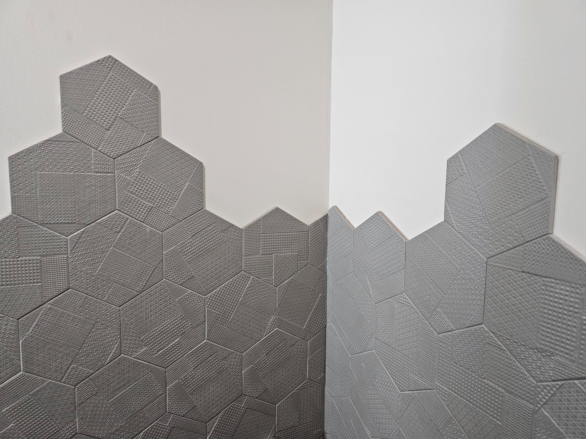 The tile used on his bathroom is an unterminated or hexagonal tile with an embossed texture. Unlike most wall tile installations which terminate all the way up at the corner of the joint between the wall plain or have a termination strip, this tile is installed with a raw staggered edge. Sometimes an edge like this is called a floating edge. If you look in the picture below, you can see that the tile at the vertical inside corner of the wall is cut to fit the shape provided by the wall plane. The top of the tile though is left raw to just follow a staggered edge of the hexagonal tile itself. The staggered unterminated edge of the tile doesn't necessarily look bad, in this example. They've been careful during installation to not let mortar ooze out in a way that looks rough or unfinished. At the lower tiles though, in the picture below, you can see the top of that unterminated edge. There are two problems with that kind of unterminated edge. First, you can see the raw edge of the tile itself. Since this is not a solid piece of natural material, the front or exposed face of the tile is a different color than the internal composition or body of the tile. When it's exposed like this and doesn't have a terminating edge you can see that difference which makes it visually apparent that it's not a real material. It's a manufactured material. The second problem with this type of unterminated edge is that the mortar itself has a thickness of roughly 1/8 of an inch. When applied with a 3/16 inch Notch trowel, the mortar becomes compressed but still even when flattened as much as possible there still is a thickness of that mortar. Once it is dry and cured, you can still see the thickness of that mortar. In almost all cases of tile installation, unlike stone masonry or even brick masonry, the tile mortar is not intended to be exposed at the joints. In between joints, in the field of the tile, the mortar will be covered with a colored grout. In this case, the tiles are set so tightly that there is almost no visible grout line between them. The grout used is a color that's very similar to the tile and the effect works. In some cases when the tile has a variation of significant color difference between the ranges of colors within the tile it's difficult to have a grout that complements or blends. Here though because the colors continuous across the face of the tile and the grout has been matched so well, you can't even hardly notice it. You can see that joint or between the back of the tile and the wall very clearly in the next picture below. You can see there's a very thin line with a black dark shadow between the tile and the wall. In some cases, when a tile is left unterminated at the edge, a gun applied caulking can be applied across the edge, smoothed, to almost make it look intentional. Sometimes in bathrooms without really good ventilation, if there's steam from hot water, that steam can condensate and build on the wall. When the vapor droplets accumulate enough they'll roll down the wall and in this case they'll sit on top of and get back behind the edge of the tile. That's one of the downsides, here though, this bathroom is just like a powder room with a toilet and a sink, but it doesn't have a shower. Most of the steam moisture buildup in bathrooms comes from showers. In the picture below you can see the distance between the other side of the ceiling and the top of the tile. Here they were at least smart enough to bring the top of the tile to a point that's above eye level. Typical eye levels are around 60 to 75 inches above the finished floor height. The mirror used above the bathroom sink has an integral LED light that wraps the perimeter of the round mirror. It basically provides a dramatic high contrast to the adjacent tile. That high contrast accentuates the texture of the tile. This tile has this intentional embossed or relief texture which is well employed when lit in a way that emphasizes the texture. The next picture below shows a close-up view of that edge at the top of the tile. Dupont Kitchen & Bath Can Help As dedicated and local DC metropolitan kitchen and bath builders and installers, we pride ourselves on turning your visions into reality. Your kitchen and bathrooms can reflect your unique style and be designed with personalized aesthetics. It can be a process from creative designs to the realization of your dream space. Specializing in kitchen and bath construction, we bring expertise and skills to every project. If you're contemplating a renovation, upgrade, or modernization in the local market, we're happy to be your team. Consult with us, and we can start together on a path to redefine and elevate your DC living experience. You can visit our company website at https://dupontkitchenbathdc.com/ Here at our website you will find a simple and convenient web form that you can fill to contact us quickly.